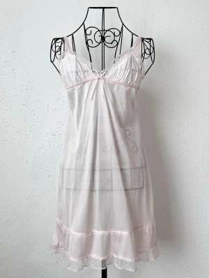 Ljusrosa spetsnattlinne med rosett - Sött ljusrosa nattlinne med tunna axelband, spetsdetaljer och liten rosett framtill. Lite genomskinlig. Passar storlek: 32-36. Dockans storlek: 34.