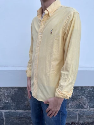 Ralph Lauren skjorta - Klassisk gul skjorta från Ralph Lauren med button-down krage och broderad logga på bröstet. Skjortan har lång ärm, normal passform och är tillverkad i mjuk bomull. Perfekt för en avslappnad men ändå stilren look.