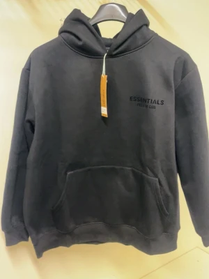 Svart Essentials hoodie - Snygg svart hoodie från Essentials Fear of God med tryck på bröstet och ryggen. Klassisk känguruficka framtill och stor huva. Tillverkad i mjukt material som känns skönt mot huden. Perfekt för en avslappnad och trendig streetwear-look.