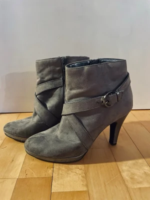 Gråa boots med klack och spänne - Snygga gråa boots med hög klack och dekorativa remmar med metallspänne. Dragkedja på insidan för enkel påtagning. Tillverkade i mockaimitation som ger en mjuk och stilren look. Perfekta för dig som vill ha en trendig och elegant stil. Står att det är storlek 40 men skulle säga att den är mer 38/39. Väl använda men ser fortfarande snygga ut!