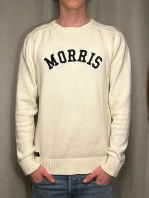 Vit stickad tröja från Morris - Vit stickad tröja från Morris med svart textlogga på bröstet. Tröjan har rund halsringning, ribbade muddar vid ärmslut och nederkant samt en klassisk, rak passform. Perfekt för en stilren och avslappnad look.