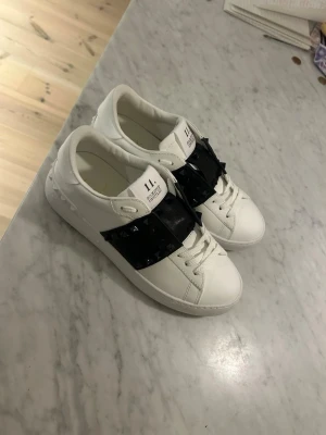 Vita sneakers med svarta nitar från Valentino - Säljer ett par vita sneakers från Valentino med bred svart rem över vristen dekorerad med svarta nitar. Skorna har klassisk låg profil, snörning och är tillverkade i skinn. Perfekta för dig som vill ha en edgy och stilren look.
