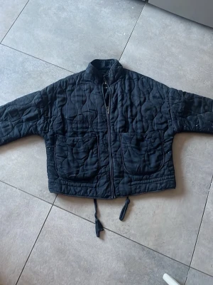 Svart quiltad jacka från Zara - Svart quiltad jacka från Zara med hög krage, dragkedja framtill och två stora fickor. Jackan har dragsko i nederkant och ett vågigt mönster i tyget. Materialet är mjukt och vadderat, perfekt för lager på lager under kyligare dagar.