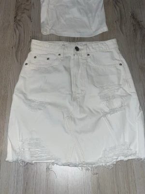 Vit jeanskjol med slitningar - Snygg vit kort jeanskjol med slitna detaljer och rå kant nertill. Kjolen har fem fickor, knapp och dragkedja framtill. Perfekt för en trendig och avslappnad look. + man får ett linne med på köpet!