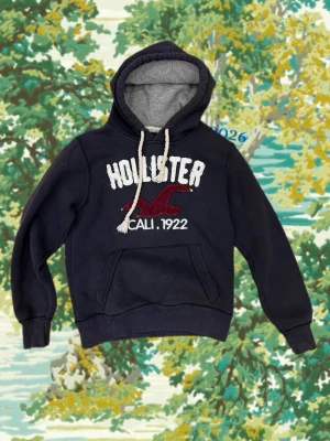  Hollister hoodie  - Mörkblå hoodie från Hollister med stor vit logga och röd fågel på bröstet samt texten 'CALI. 1922'. Utrustad med huva, vita dragsnören och magficka. Insidan av huvan är grå. Perfekt för en avslappnad och cool stil.