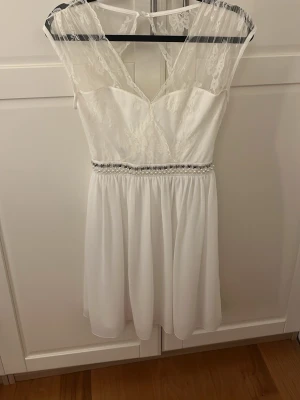 Vit spetsklänning med pärldetaljer - Elegant vit klänning med spets upptill och v-ringning både fram och bak. Midjan är dekorerad med pärlor och små stenar för extra detalj. Kjolen är lätt och faller mjukt. Perfekt för student eller sommarfest.