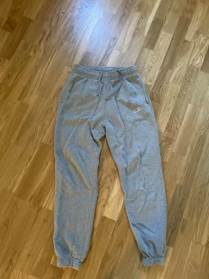 Gråa mjukisbyxor från Nike - Säljer ett par klassiska gråa mjukisbyxor från Nike med dragsko i midjan och resår vid bensluten. Byxorna har sidofickor och en liten Nike-logga broderad på vänster ben. Perfekta för chill dagar eller träning.