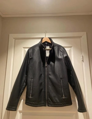 Svart skinnjacka från Hampton republik  - Stilren svart skinnjacka från Hampton republik med rak passform och ståkrage. Jackan har dragkedja framtill, två sidofickor med dragkedja och mjukt foder i kragen för extra komfort. Perfekt för dig som vill ha en klassisk look med edge.