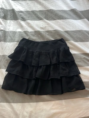 Svart volangkjol med resårmidja - Säljer en svart kortkjol med flera lager volanger och bred resår i midjan. Kjolen har en luftig och flowy känsla, perfekt för att styla med en enkel topp eller hoodie. Materialet känns lätt och bekvämt.