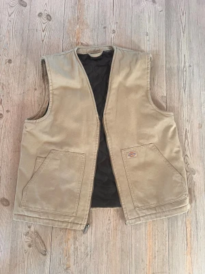 Beige arbetsväst från Dickies - Säljer en beige arbetsväst från Dickies med två stora framfickor och quiltat svart innerfoder. Västen har rak passform och dragkedja framtill. Perfekt för lager på lager och streetwear-stil.