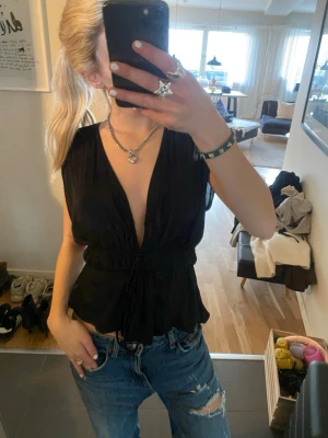 Svart omlottopp med snörning - Säljer en svart omlottopp med djup v-ringning och snörning i midjan. Toppen är ärmlös och har en avslappnad passform som ger en snygg siluett. Perfekt att styla med jeans eller kjol för en trendig look.