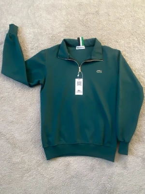 Grön ziptröja från Lacoste - Mörkgrön ziptröja från Lacoste med halv dragkedja och ståkrage. Tröjan har ribbade muddar vid ärmslut och nederkant samt den klassiska krokodilloggan broderad på bröstet. Materialet är mjukt och bekvämt, perfekt för en avslappnad stil.