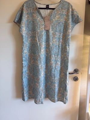 Blåvit kortklänning oanvänd  - Kortklänning i blått och vitt med ett elegant blommigt mönster. Klänningen har korta ärmar, v-ringning och rak passform. Tillverkad i ett lätt och luftigt material, perfekt för varma dagar. Oanvänd.