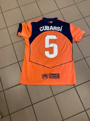 Orange FC Barcelona matchtröja Nike - Säljer en orange FC Barcelona matchtröja från Nike med mörkblå detaljer och tryck. Tröjan har korta ärmar, nummer 5 och namnet Cubarsí på ryggen samt sponsorloggor från Spotify och UNHCR. Perfekt för fotbollsträning eller att bära som supporter.