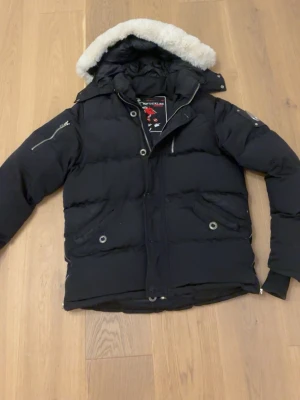 Svart dunjacka med pälshuva Moose Knuckles - Säljer en svart dunjacka från Moose Knuckles med vit päls . Jackan har flera fickor med dragkedja och tryckknappar samt den ikoniska logotypen i metall på ärmen. Perfekt för kalla vinterdagar. metall loggan på sidan är dock lite repad och saken man hänger upp jackan med har lossnat