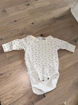Vit body med blommönster från Zara -  Söt vit långärmad body från Zara med små blommor i grönt och rosa. Bodyn har omlottkrage och tryckknappar i grenen för enkel påklädning. Perfekt för dig som gillar gulliga och stilrena babykläder.