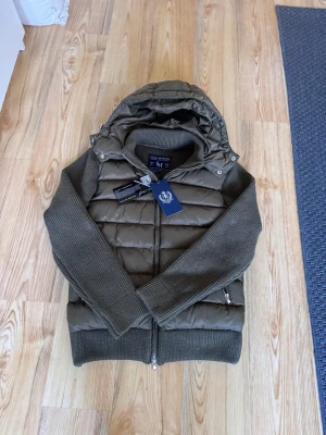 Leon Marcel Hybrid Puffer Jacka – Olive | Ny Med Prislapp - Ny med prislapp — Leon Marcel hybridjacka med quiltad pufferkropp och stickade ärmar. En unik Skandinavisk design du inte hittar någon annanstans. Avtagbar huva och lyxiga microfleecefickor som håller händerna varma — detaljer som gör skillnad. Premium kvalitet, mångsidig olivfärg som passar till allt. Perfekt för höst/vinter. Säljer direkt för att erbjuda ett bättre pris än butik. 📦 Spårad frakt, väl förpackad.