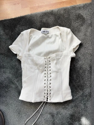 Vit ribbad korsettopp från I.AM.GIA - Trendig vit ribbad korsettopp från I.AM.GIA med snörning framtill och korta ärmar. Toppen har fyrkantig urringning och markerad midja för en snygg siluett. Liten defekt på baksidan längst ner- använd ett fåtal gånger 
