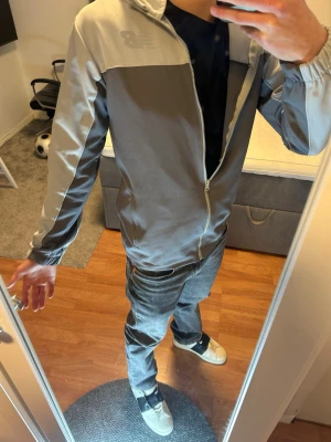 Grå vindjacka från New Balance - Snygg grå vindjacka från New Balance med dragkedja framtill och elastiska muddar vid ärmsluten. Jackan har två sidofickor och en sportig design med ljusare partier på axlar och ärmar. Perfekt för dig som gillar streetwear och enkel stil.