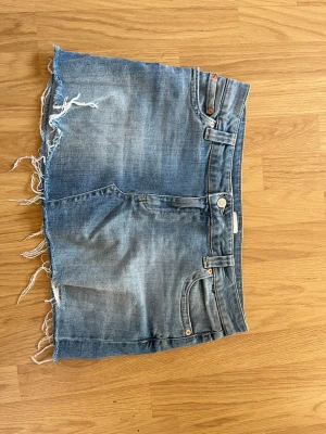 Blå jeanskjol med fransig kant - Säljer en blå jeanskjol i klassisk denim med rå, fransig kant nertill. Kjolen har fem fickor, bälteshällor och knappgylf. Perfekt för dig som gillar en avslappnad och trendig stil. Kjolen är i storlek 40 från Nelly och nypris är 500 kr.