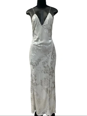 Ljusgrå långklänning med silvrigt mönster - Klänning från Zara. Tunt satinliknande material med silvermönster i tyget. Tunna axelband, v-ringning fram och öppen rygg.