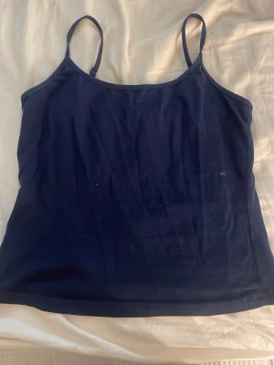 Mörkblå linnetopp med smala band - En enkel och stilren mörkblå linnetopp med tunna axelband. Toppen har en rak passform och är tillverkad i ett mjukt, stretchigt material som känns bekvämt mot huden. Perfekt att matcha med jeans eller kjol för en clean look.
