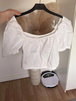 Vit croppad offshoulder topp - Söt vit croppad topp med offshoulder-design och korta puffärmar. Tillverkad i bomull med resår vid axlar och midja för en bekväm passform. Perfekt till jeans eller kjol för en trendig look.