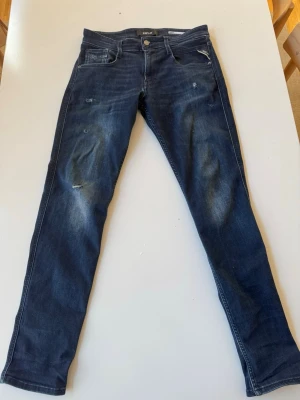 Mörkblå skinny jeans från Replay - Snygga mörkblå skinny jeans från Replay i storlek 31 med slitningar och lätt tvättade detaljer på benen. Klassisk femficksmodell med dragkedja och knapp. Jeansen har en modern passform och är tillverkade i stretchigt denimtyg för extra komfort