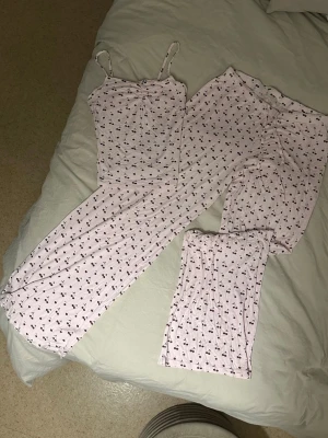 Ljusrosa pyjamas med körsbärsmönster - Supersöt ljusrosa pyjamas med körsbärsmönster. Setet består av ett linne med smala axelband och långa byxor i mjukt, stretchigt material. Perfekt för mysiga kvällar hemma. Materialet känns lätt och luftigt mot huden.