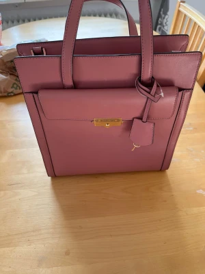 Rosa handväska från Michael Kors - Snygg rosa handväska från Michael Kors i stilren boxig modell. Väskan är gjord i slätt läder med guldfärgade metalldetaljer och har både handtag och axelrem. Invändigt finns fack med dragkedja och logotypmönstrat foder.
