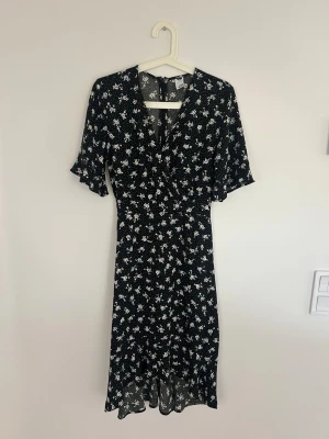 Svart blommig klänning  - Svart långklänning med vitt blommönster och knytning bak . Klänningen har v-ringning, halvlånga ärmar och slits nertill. Perfekt för dig som gillar en feminin och avslappnad stil.