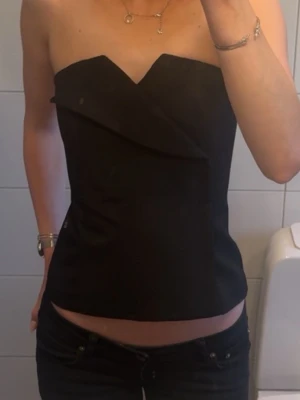 Svart axelbandslös topp med volang - Snygg svart axelbandslös topp med unik volangdetalj framtill. Toppen har en croppad passform och är perfekt för dig som vill sticka ut. Tillverkad i ett slätt material som ger en stilren look.