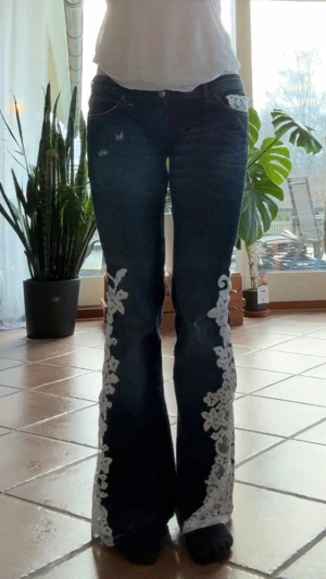 Mörkblå bootcut jeans med spets - Unika mörkblå bootcut jeans med vita spetsapplikationer längs benen och fickdetalj fram. Jeansen har låg midja och klassisk femficksmodell. Jag har sytt på spetsen själv då jeansen blir unika. Jag har även sytt jeansen lågmidjade. Vid någon fundering är det bara att fråga!