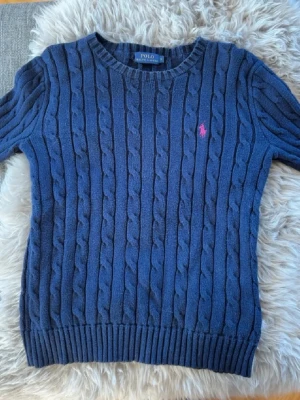 Polo Ralph Lauren - Snygg mörkblå kabelstickad tröja från Polo Ralph Lauren med rund halsringning och rosa broderad logga på bröstet. Tröjan har ribbade muddar vid ärmslut och nederkant. Perfekt för dig som gillar klassisk och tidlös stil. Nyskick och super fin!!