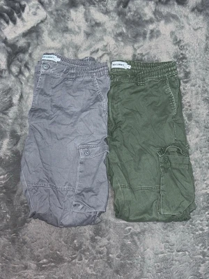 2 New garments cargobyxor i ett - Snygga cargobyxor i grönt och grått från New Comfort. Byxorna har flera praktiska fickor med lock på sidorna och elastisk midja för skön passform. Tillverkade i ett mjukt bomullstyg som ger en avslappnad look. Perfekta för en casual streetstyle.