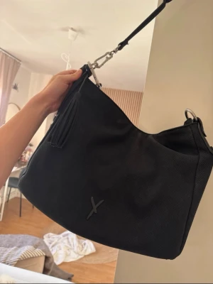 Svart axelväska från Suri Frey - Stilren svart handväska från suri frey med mjuk, rundad form och avtagbar axelrem. Väskan har en dekorativ tofs och ett diskret Y-logotyp framtill. Tillverkad i slätt konstläder med silvriga metalldetaljer. Perfekt för dig som gillar minimalistisk design.