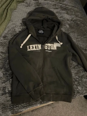 Mörkgrå hoodie från Lexington - Mörkgrå hoodie från Lexington med vit logga på bröstet och vita snören i huvan. Klassisk zip hoodie med fickor framtill och mjukt material som känns skönt mot huden. Perfekt för chill dagar eller när du vill ha en avslappnad stil.