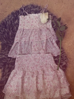 Gulligt blommigt set i rosa – topp & kjol - Detta är ett sött och somrigt set i ljusrosa toner med ett småblommigt mönster. Tyget är lätt och luftigt!🌸Toppen matchar kjolen med samma blommiga mönster och smockade överdel. Den har tunna axelband och en lätt volang nedtill!🌸🌸
