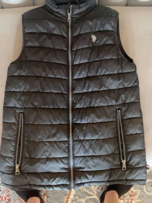Svart dunväst från U.S. Polo Assn. - Snygg svart dunväst från U.S. Polo Assn. med quiltad design och två stora dragkedjefickor framtill. Västen har hög krage och en liten logga på bröstet. Perfekt för lager på lager och ger en sportig vibe.