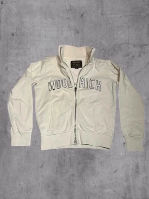 Vit zip hoodie från Woolrich - Snygg vit hoodie från Woolrich med dragkedja framtill och broderad logga i svart på bröstet. Tröjan har hög krage, två fickor och ribbade muddar. Tillverkad i mjuk bomullsblandning som känns skön mot huden. Perfekt för en avslappnad streetwear-look.