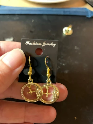 Guldörhängen med måne och stjärna - Säljer ett par snygga guldörhängen .Örhängena har en rund form med en måne och ett ansikte i relief samt en hängande stjärna i mitten. Ena sidan av cirkeln är dekorerad med små glittrande stenar för extra bling.