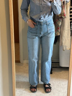 Ljusblå raka jeans med fickor - Säljer ett par ljusblå jeans med raka ben och hög midja. Jeansen har stora framfickor och en bakficka med knappdetalj. Klassisk femficksmodell i mjukt denimtyg, perfekta till vardagslooken.