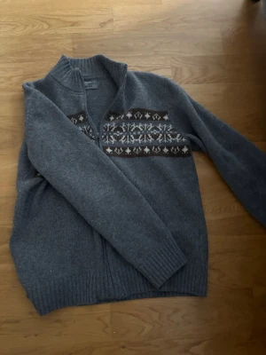 Fet stickad tröja  - Mysig grå stickad kofta med dragkedja och högt ribbad krage. Har ett klassiskt mönster i vitt och brunt över bröstet. Långa ärmar och ribbade muddar vid ärmslut och nederkant. Perfekt för kalla dagar.