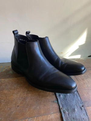 Svarta Chelsea boots i läder - Stilrena svarta Chelsea boots i slätt läder med klassisk resår på sidorna och dragflikar bak. Snygg minimalistisk design som passar till många outfits. Perfekta för dig som gillar tidlös och enkel stil.
