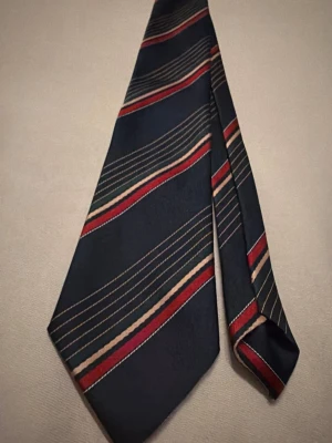 Mörkblå slips med ränder - Snygg mörkblå slips med diagonala ränder i rött, beige och guld. Tillverkad i ett glansigt tyg som ger en elegant känsla. Perfekt för att lyfta din outfit med lite färg och stil.