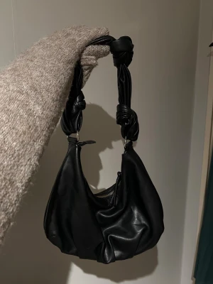 Svart axelväska med knutdetaljer - Trendig svart handväska i mjukt fejkläder med unik axelrem dekorerad med stora knutar. Väskan har en rundad form och stängs med dragkedja. Perfekt för dig som gillar en edgy och modern stil.