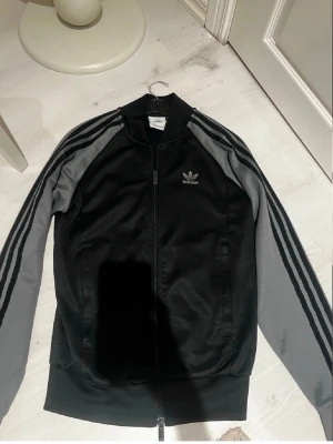 Adidas hoodie - Snygg svart ziptröja från Adidas med grå ärmar och klassiska tre ränder längs ärmarna. Tröjan har dragkedja framtill, ståkrage och Adidas logga på bröstet. Perfekt för en sportig och avslappnad stil.