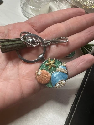 Nyckelring/väsksmycke  med havstema och tofs - Säljer en unik nyckelring / väsksmycke med havstema, dekorerad med snäcka, sjöstjärna, blå snäcka och pärlor på en grön bakgrund. Metallring och karbinhake i silverfärg samt en mörkgrön tofs i mockaimitation. Perfekt accessoar för dig som gillar strandvibbar!