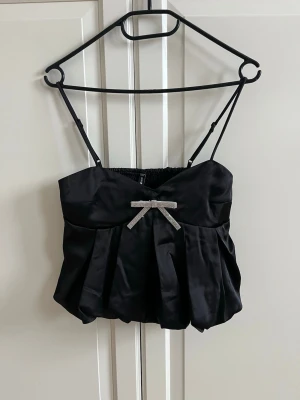 Satintopp - Superfin svart Volangtopp i satin från new yorker. Man får med en tillbehörande silvrig diamantrosett som är avtagbar. Nyskick. Storlek xxs, men passar mig som vanligtvis har s och xs i toppar🩷
