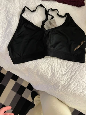 Svart sport-bh från Newbody - Sportig svart bh från Newbody med bred resår under bysten och justerbara axelband. Tillverkad i ett mjukt och stretchigt material som ger bra stöd. Diskret logga i guld på sidan. Perfekt för träning eller vardag.